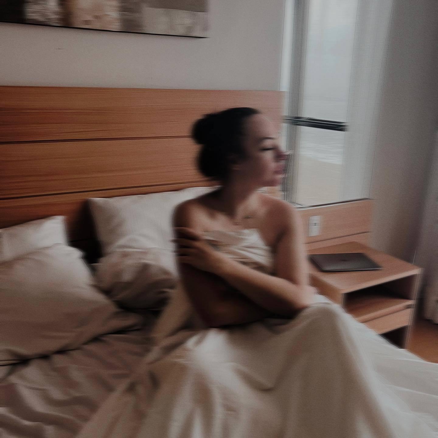 Слегка размазанная фотография Синди в кровати под одеялом с обнажёнными руками и плечами.