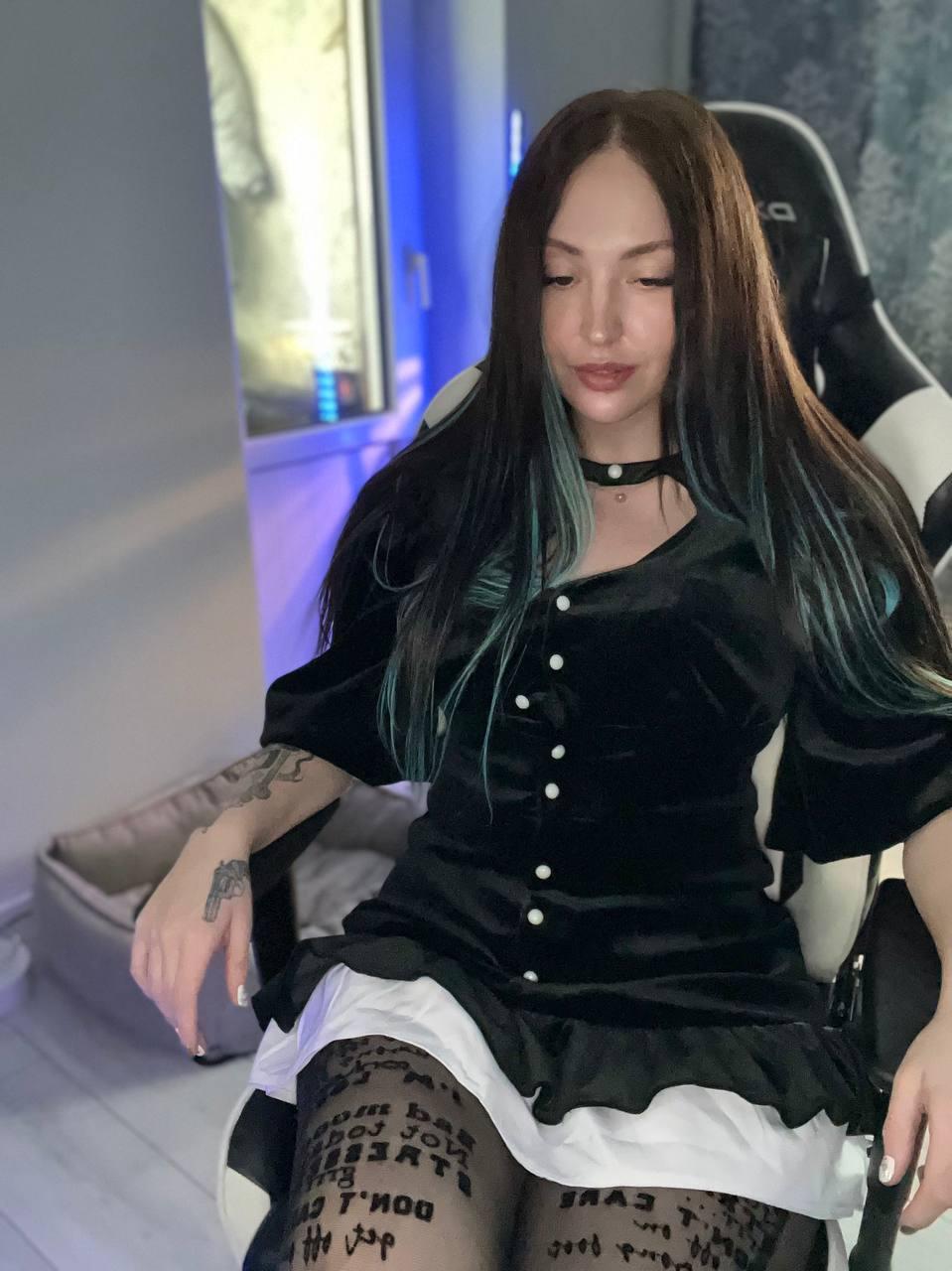 Красотка Sindi в игровом кресле в коротком платье и чёрных чулках.